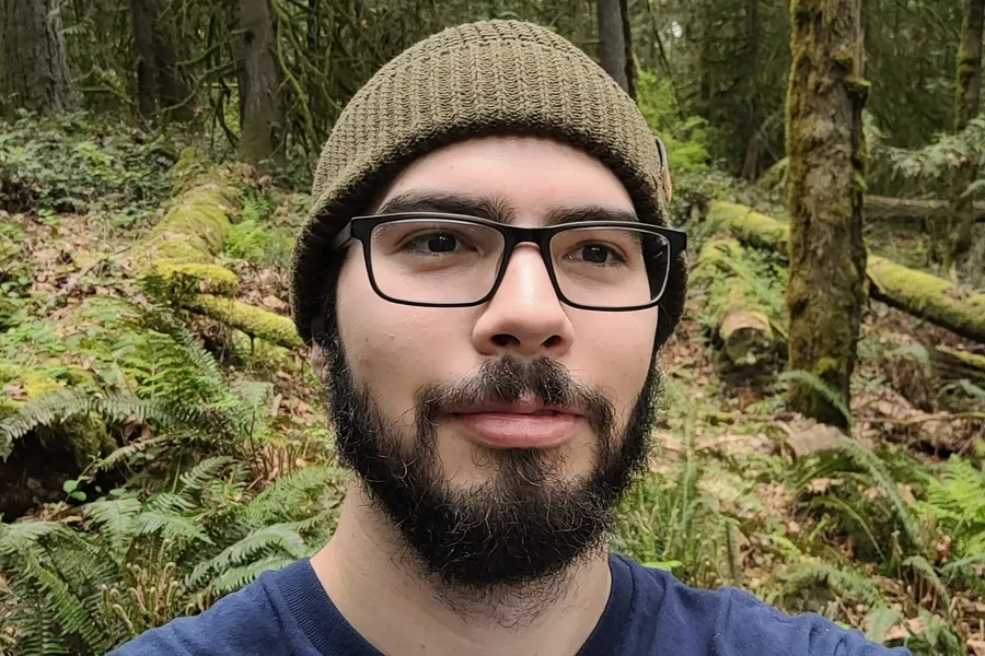 Eric Chavez in a beanie and glasses, standing in a mossy forest of ferns and fallen logs